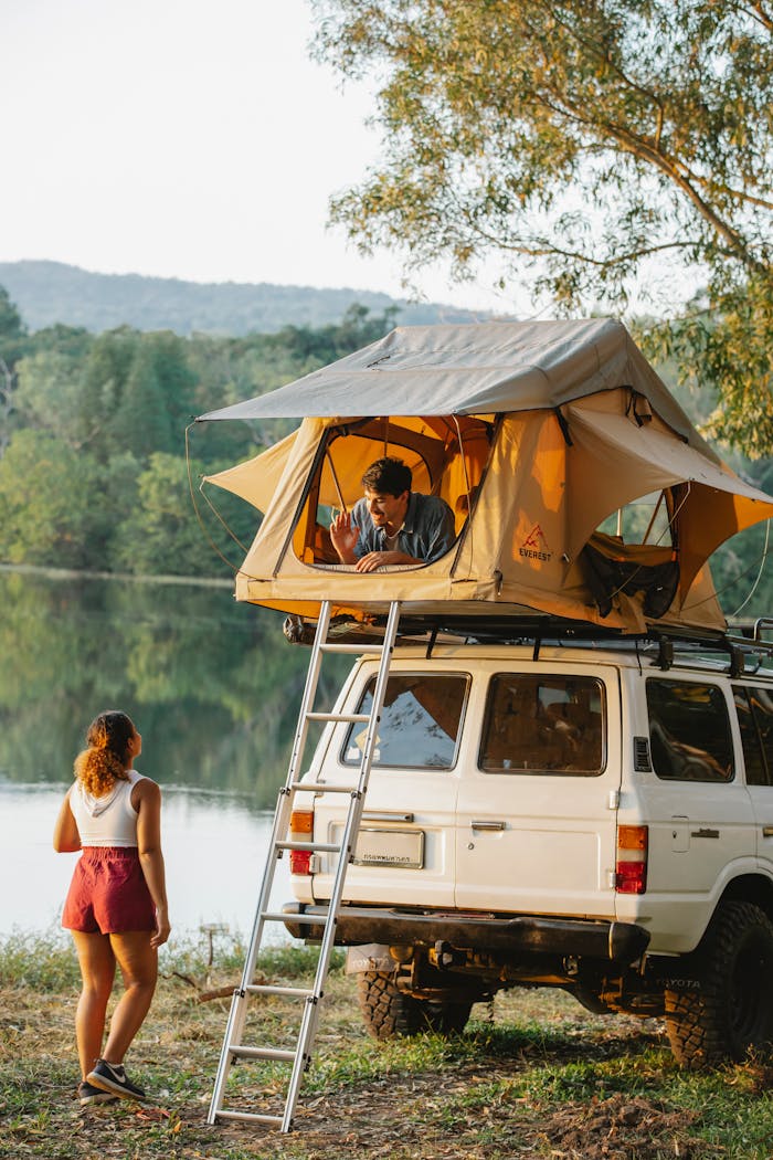 about-01 Back view of unrecognizable young female traveler standing on lake shore and talking to boyfriend resting in tent placed on SUV car roof